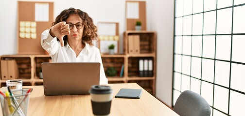 Middle age hispanic woman working at the office wearing glasses looking unhappy and angry showing rejection and negative with thumbs down gesture. bad expression.