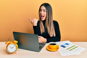 Young hispanic woman working at the office with laptop pointing thumb up to the side smiling happy with open mouth
