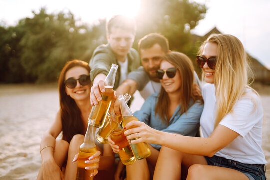 Group of friends cheers and drink beers on the beach. Young friends relaxing on the beath having picnic, toasting with beerus. People, lifestyle, travel, nature and vacations concept.
