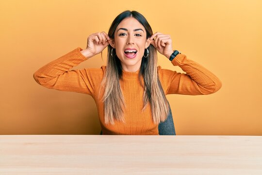 Young hispanic woman wearing casual clothes sitting on the table smiling pulling ears with fingers, funny gesture. audition problem