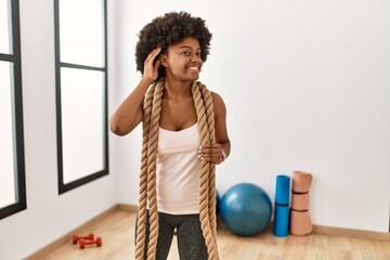 Young african american woman with afro hair at the gym training with battle ropes smiling with hand over ear listening an hearing to rumor or gossip. deafness concept.