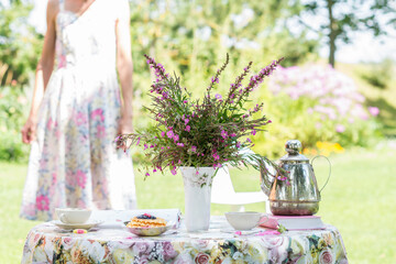 Table with flowery tablecloth and cup of tea, wild flowers bouquet of  rosebay willowherbs or  fireweed in a vase , cake and teapot , woman standing in background outdoors in summer time in the garden