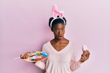 Young african american girl wearing cute easter bunny ears painting egg in shock face, looking...