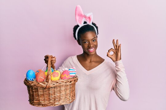 Young african american girl wearing cute easter bunny ears holding basket with painted eggs doing ok sign with fingers, smiling friendly gesturing excellent symbol