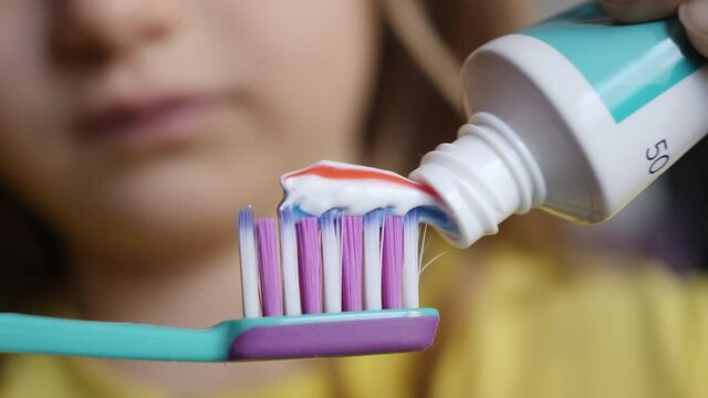 Little Girl Extruded Toothbrush With Bright Striped Toothpaste. Macro Shot Red and Blue Stripe Toothpaste Extruded Child Onto Toothbrush Closeup. Dental Health Oral Hygiene. Morning Teeth Brushing.