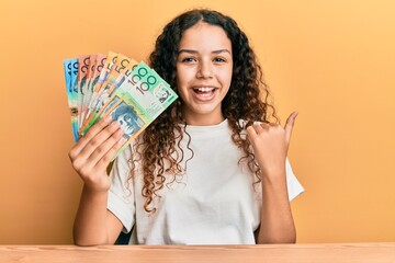 Teenager hispanic girl holding australian dollars pointing thumb up to the side smiling happy with open mouth