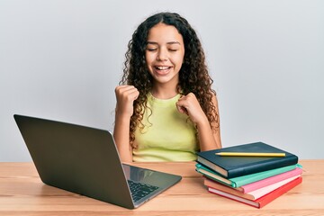 Teenager hispanic girl sitting on the table studying for school celebrating surprised and amazed for success with arms raised and eyes closed