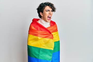 Young man wearing woman make up wrapped in rainbow lgbtq flag angry and mad screaming frustrated...