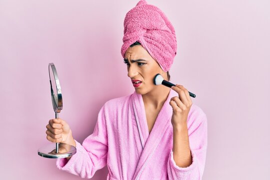 Young man wearing woman make up wearing pink robe applying blush in shock face, looking skeptical and sarcastic, surprised with open mouth