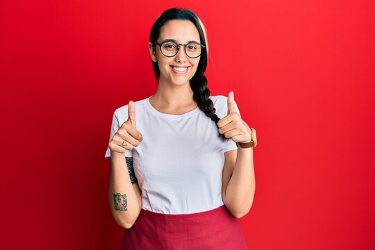 Young hispanic woman wearing professional waitress apron success sign doing positive gesture with hand, thumbs up smiling and happy. cheerful expression and winner gesture.