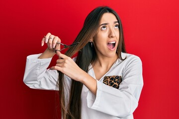 Young hispanic girl cutting hair using scissors angry and mad screaming frustrated and furious, shouting with anger looking up.