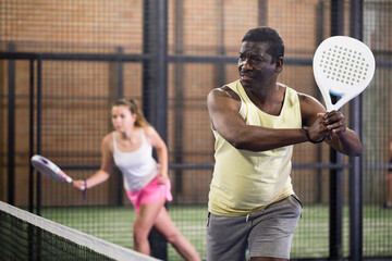 Confident African American man playing padel tennis on indoor court, ready to hit two handed...