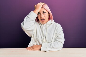 Hispanic woman with pink hair wearing casual sweatshirt sitting on the table surprised with hand on...