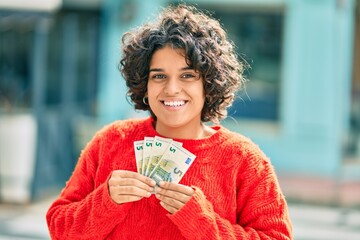 Young hispanic woman smiling happy holding 5 euro banknotes at the city.