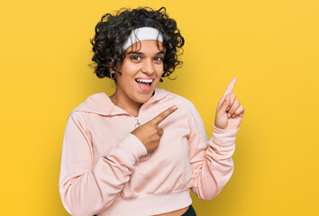 Young hispanic woman with curly hair wearing sportswear smiling and looking at the camera pointing with two hands and fingers to the side.