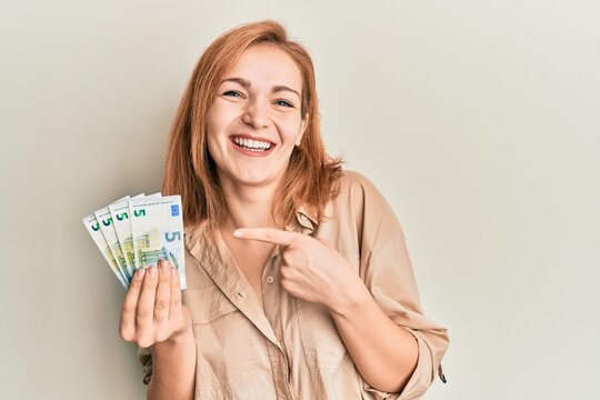Young caucasian woman holding bunch of 5 euro banknotes smiling happy pointing with hand and finger