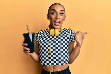 Hispanic man wearing make up and long hair wearing headphones and drinking take away soda pointing thumb up to the side smiling happy with open mouth