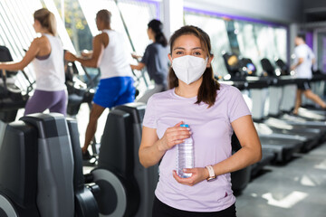 Portrait of young woman wearing protective face mask resting after workout in fitness gym holding...