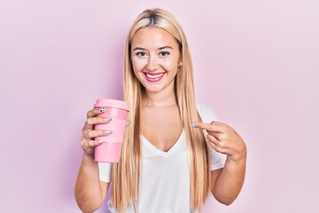Young blonde girl holding coffee smiling happy pointing with hand and finger