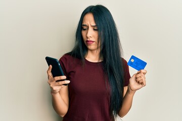 Young hispanic girl holding smartphone and credit card clueless and confused expression. doubt...
