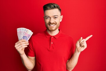 Young redhead man holding 20 polish zloty banknotes smiling happy pointing with hand and finger to the side