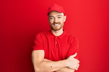 Young redhead man wearing delivery uniform and cap happy face smiling with crossed arms looking at the camera. positive person.