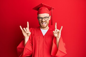 Young redhead man wearing red graduation cap and ceremony robe shouting with crazy expression doing...
