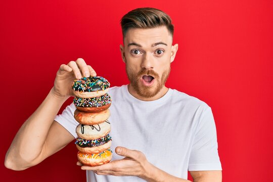 Young redhead man holding pile of tasty colorful doughnuts afraid and shocked with surprise and amazed expression, fear and excited face.