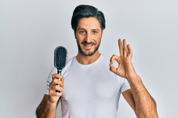 Young hispanic man styling hair using comb doing ok sign with fingers, smiling friendly gesturing...