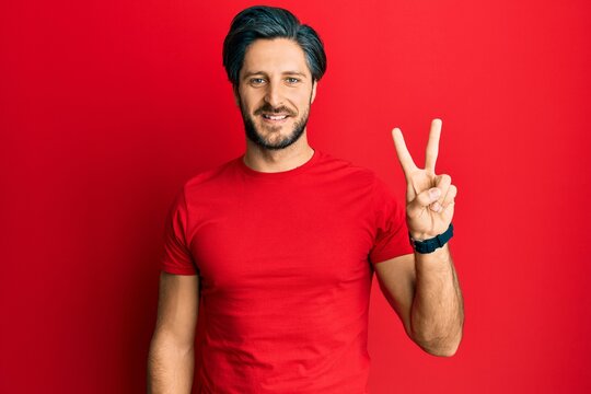 Young hispanic man wearing casual red t shirt smiling looking to the camera showing fingers doing victory sign. number two.