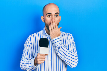 Young bald man holding reporter microphone covering mouth with hand, shocked and afraid for...