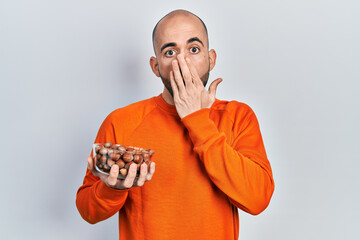 Young bald man holding raw hazelnuts covering mouth with hand, shocked and afraid for mistake....