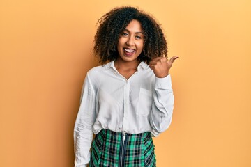 Beautiful african american woman with afro hair wearing scholar skirt smiling with happy face looking and pointing to the side with thumb up.