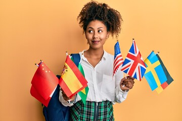Beautiful african american woman with afro hair exchange student holding countries flags smiling looking to the side and staring away thinking. - obrazy, fototapety, plakaty