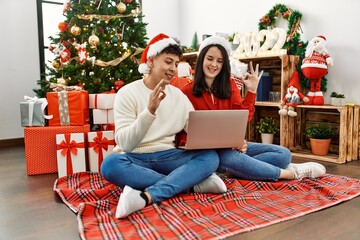Obraz na płótnie Canvas Young hispanic couple wearing christmas hat using laptop sitting by christmas tree doing ok sign with fingers, smiling friendly gesturing excellent symbol