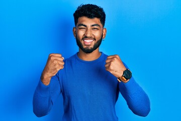 Arab man with beard wearing casual blue sweater celebrating surprised and amazed for success with arms raised and open eyes. winner concept.