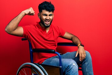 Arab man with beard sitting on wheelchair angry and mad raising fist frustrated and furious while shouting with anger. rage and aggressive concept.