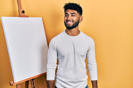 Arab man with beard standing by painter easel stand looking to side, relax profile pose with natural face and confident smile.