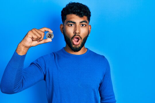 Arab man with beard holding virtual currency bitcoin scared and amazed with open mouth for surprise, disbelief face