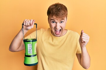 Young caucasian man holding big electric lantern screaming proud, celebrating victory and success very excited with raised arms