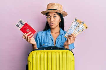 Young chinese woman holding boarding pass and passport clueless and confused expression. doubt...