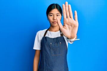 Young chinese woman wearing waiter apron doing stop sing with palm of the hand. warning expression...