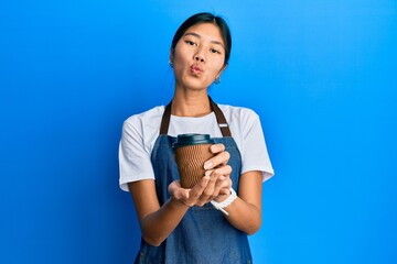 Naklejka premium Young chinese woman wearing waiter apron holding cup of coffee looking at the camera blowing a kiss being lovely and sexy. love expression.