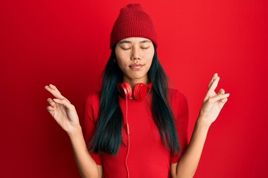 Young chinese woman listening to music using headphones gesturing finger crossed smiling with hope and eyes closed. luck and superstitious concept.