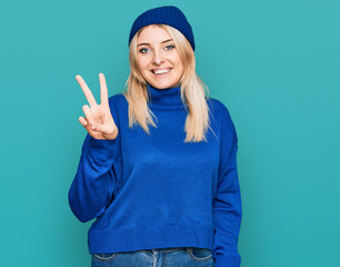 Young caucasian woman wearing wool winter sweater and cap smiling looking to the camera showing fingers doing victory sign. number two.