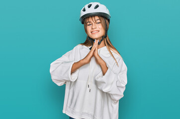Teenager caucasian girl wearing bike helmet praying with hands together asking for forgiveness smiling confident.