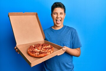 Teenager hispanic boy eating tasty pepperoni pizza sticking tongue out happy with funny expression.