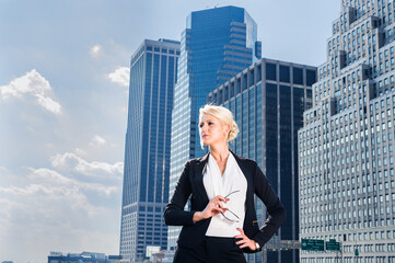 Fototapeta na wymiar Dressing in white underclothes and a black suit unbuttoned, holding glasses, a young pretty blonde businesswoman is standing in the front of a business district and hopefully looking forward..