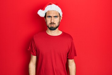 Young hispanic man wearing christmas hat puffing cheeks with funny face. mouth inflated with air,...
