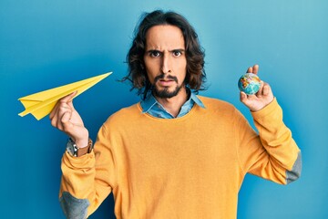Young hispanic man holding paper plane and world ball clueless and confused expression. doubt...